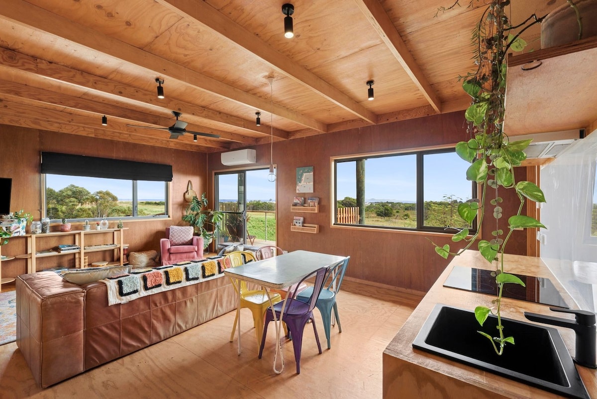 An open living space features wooden beams on the ceiling and large windows offering views of the surrounding greenery. A colorful dining table is flanked by bright chairs, while a cozy couch adorned with textured blankets creates a welcoming atmosphere. A kitchen area is visible with modern appliances.