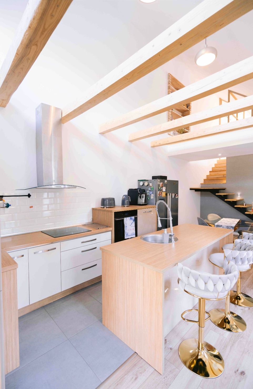 The kitchen area is characterized by modern white cabinetry and wooden accents. Stainless steel appliances complement the space, which features a central island with stylish bar stools. Light floods in through large windows, highlighting the open design and airy feel.