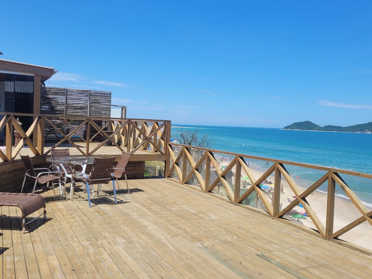 A wooden deck is showcased with a spacious area featuring outdoor chairs and tables, overlooking the beach. The vibrant blue sea and clear sky provide a scenic backdrop, while the coastline is visible in the distance.