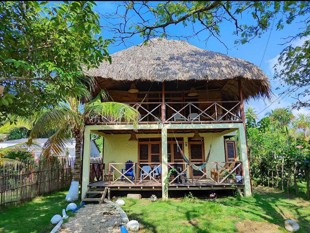 This two-story house features a thatched roof and a spacious balcony. The exterior walls are painted in soft tones, complemented by wooden accents. Hammocks are seen hanging near chairs on the porch, surrounded by lush greenery and a well-maintained garden.