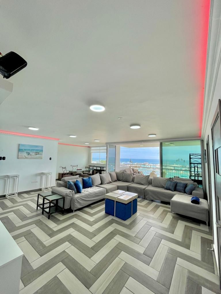 A spacious living area features a large sectional sofa adorned with blue and gray cushions. Floor-to-ceiling windows offer vibrant views of the beach, while a modern dining space is visible in the background. The floor is designed with a distinct herringbone pattern.