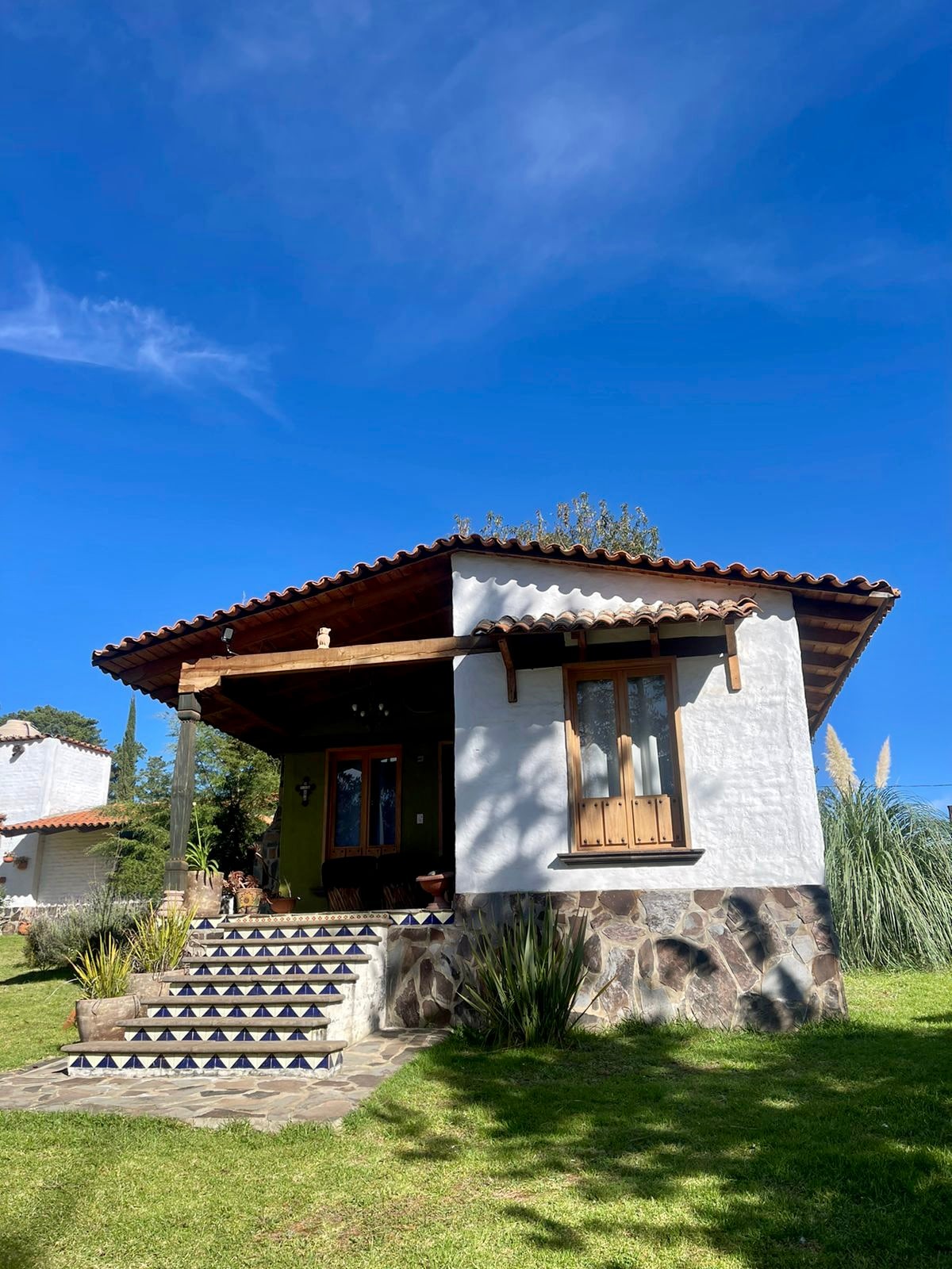 A charming cabin is set against a clear blue sky, featuring a pitched roof and wooden accents. Stone steps lead to a welcoming entrance adorned with plants and decorative elements. Lush greenery surrounds the structure, enhancing the serene outdoor space.
