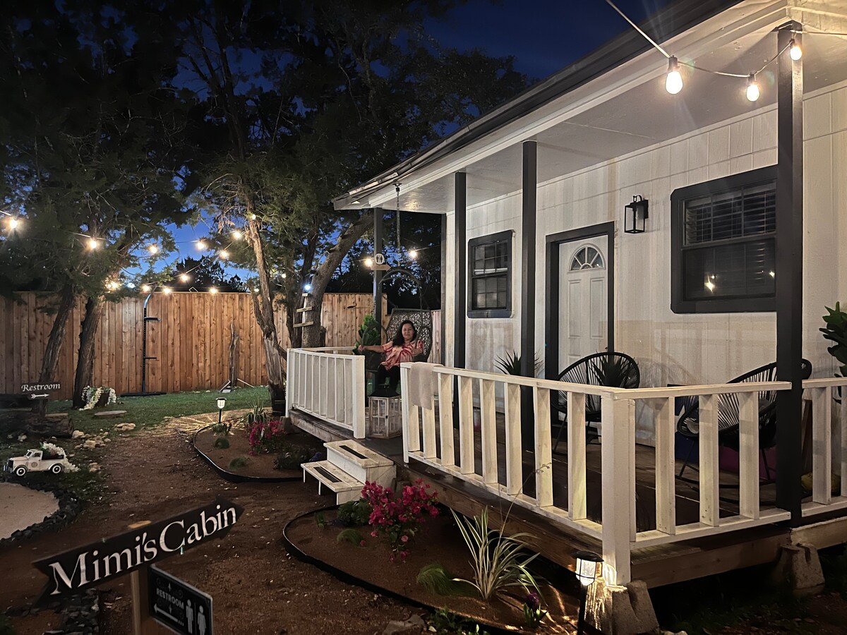 The exterior of Mimi's Cabin is depicted at night, showcasing a welcoming porch adorned with string lights. A sign reading 'Mimi's Cabin' is positioned near the entrance. The surrounding area features landscaped flora and a cozy seating arrangement, inviting relaxation in a tranquil outdoor setting.