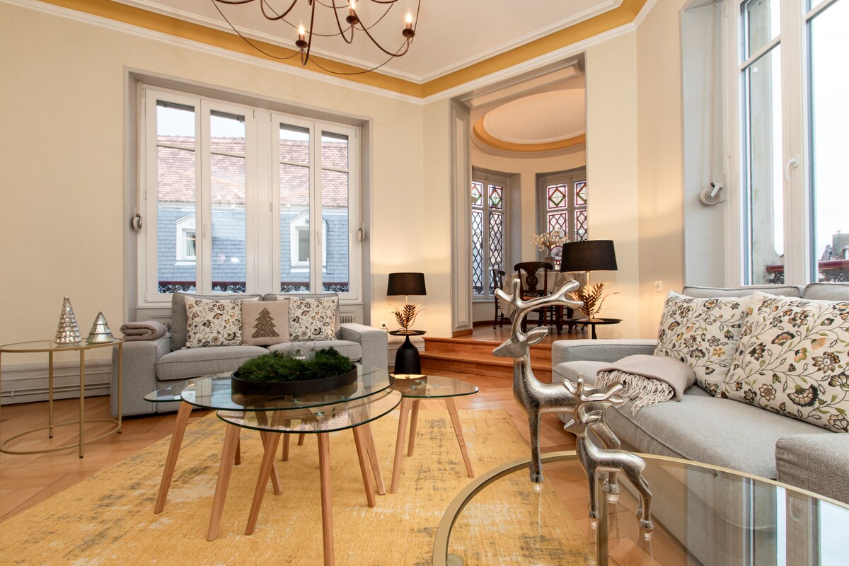 A spacious living room features large windows allowing natural light to illuminate the warm, neutral tones of the decor. Comfortable sofas are arranged around glass tables, complementing the parquet flooring. An elevated area with stained glass details enhances the character of the room.