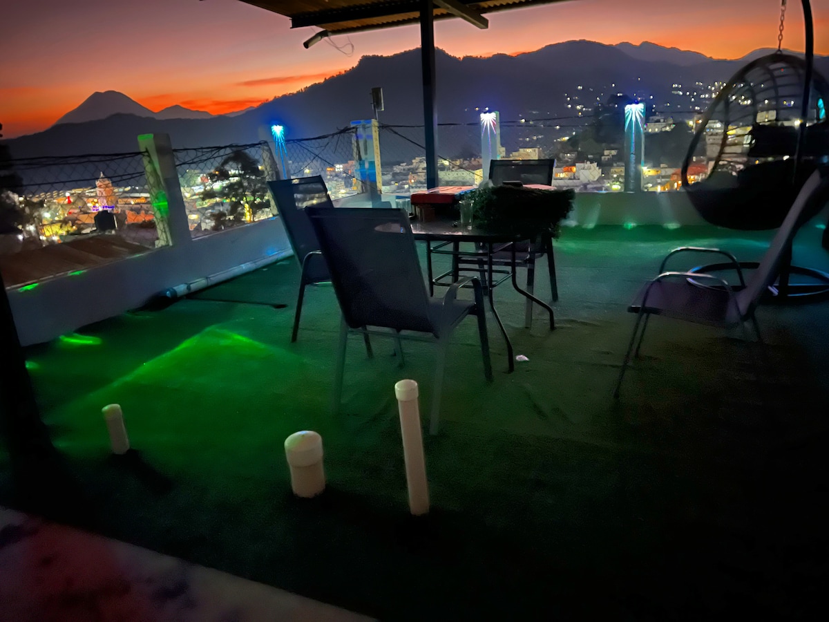 An outdoor seating area is visible on the Roof Garden, featuring a table surrounded by four chairs. The space is softly illuminated by colored lights, enhancing the view of the distant mountains under a twilight sky. A hanging chair is also included for relaxation.