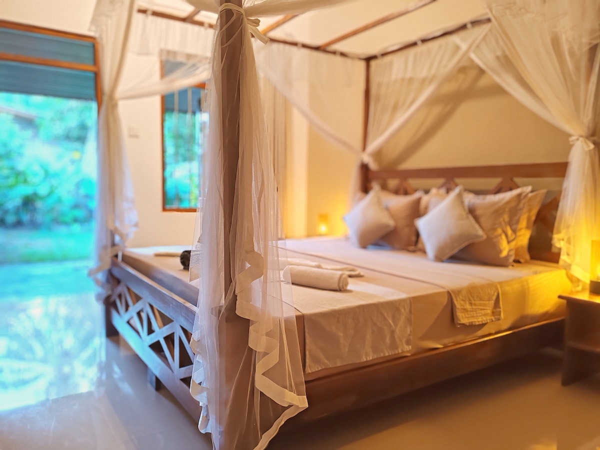 The private room features a spacious bed framed by a decorative canopy. Soft white linens are complemented by plush pillows. A serene outdoor view is visible through large windows, softly illuminated by warm lighting that enhances the calming ambiance.