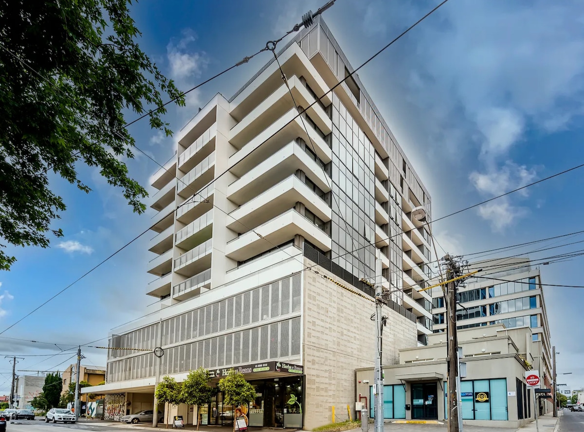 A modern multi-story apartment building is captured from an angled view. The façade features large glass panels and balconies, surrounded by greenery at the base. The busy street scene below includes shops and a tram stop, emphasizing the urban setting.