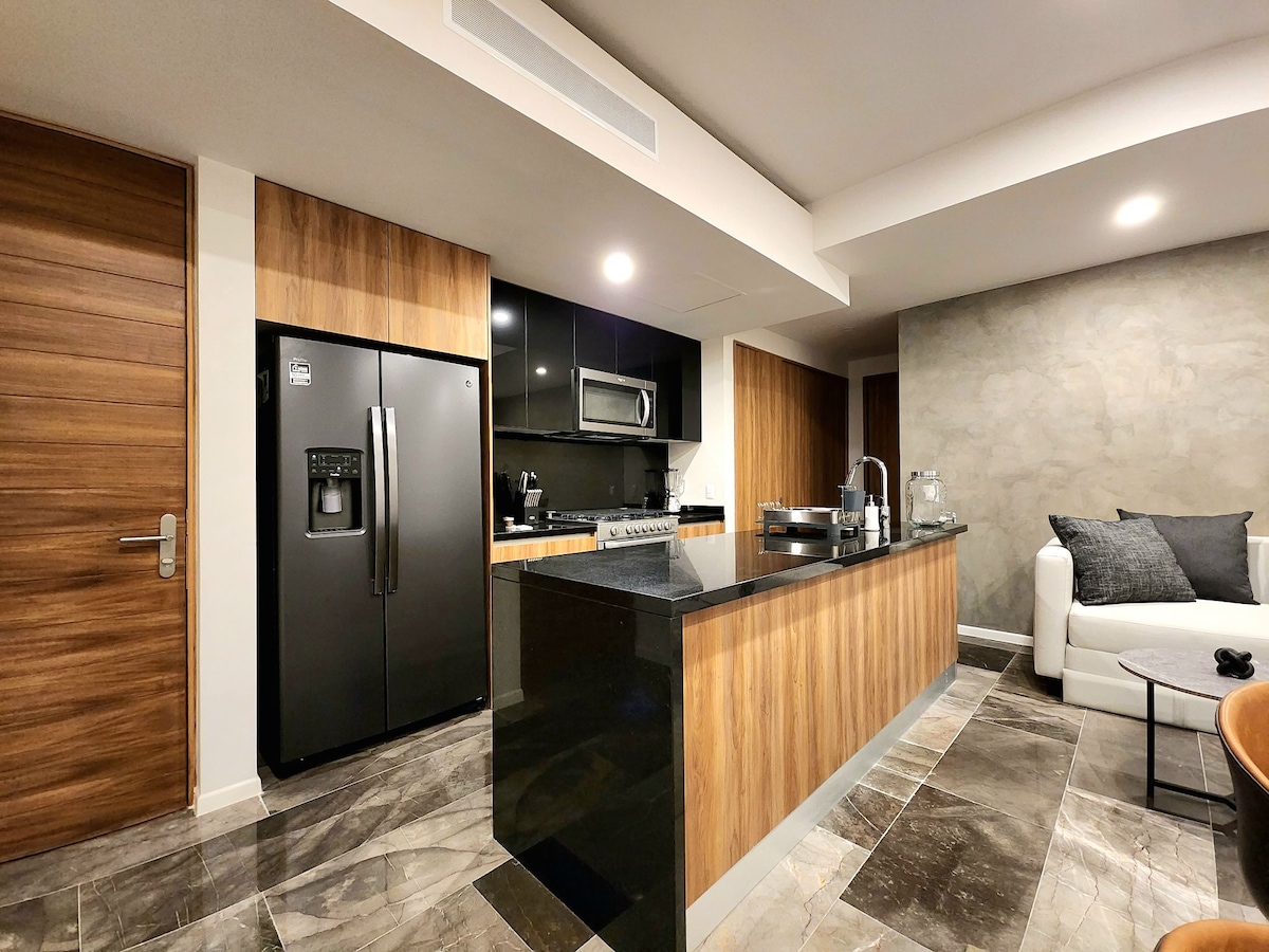 A modern kitchen is showcased with sleek cabinetry and a glossy black countertop. Stainless steel appliances are integrated, including a refrigerator and an oven. The open layout promotes a seamless connection to the living area, where a light-colored sofa is positioned against a textured wall.