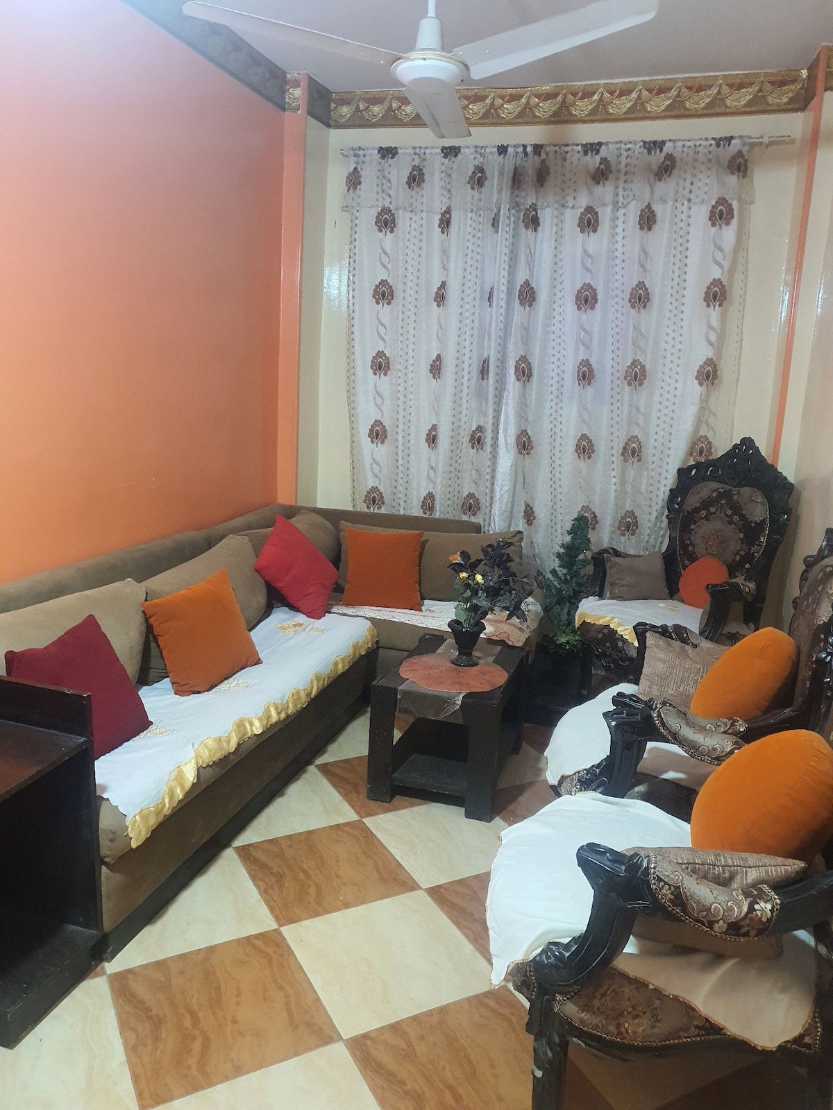 A cozy sitting area features a corner sofa adorned with colorful cushions and an elegant wooden coffee table. Two ornate armchairs frame the space, complemented by a potted plant. Natural light filters through sheer curtains, enhancing the inviting ambiance with warm orange walls.