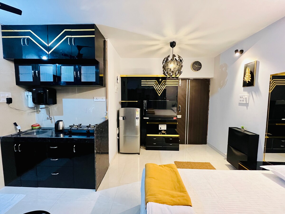 A modern studio apartment is displayed, featuring a kitchenette with black cabinets and a gas stove. A refrigerator is adjacent to the workstation area. The living area showcases a sleek design with decorative lighting and essential furnishings, enhancing functionality.