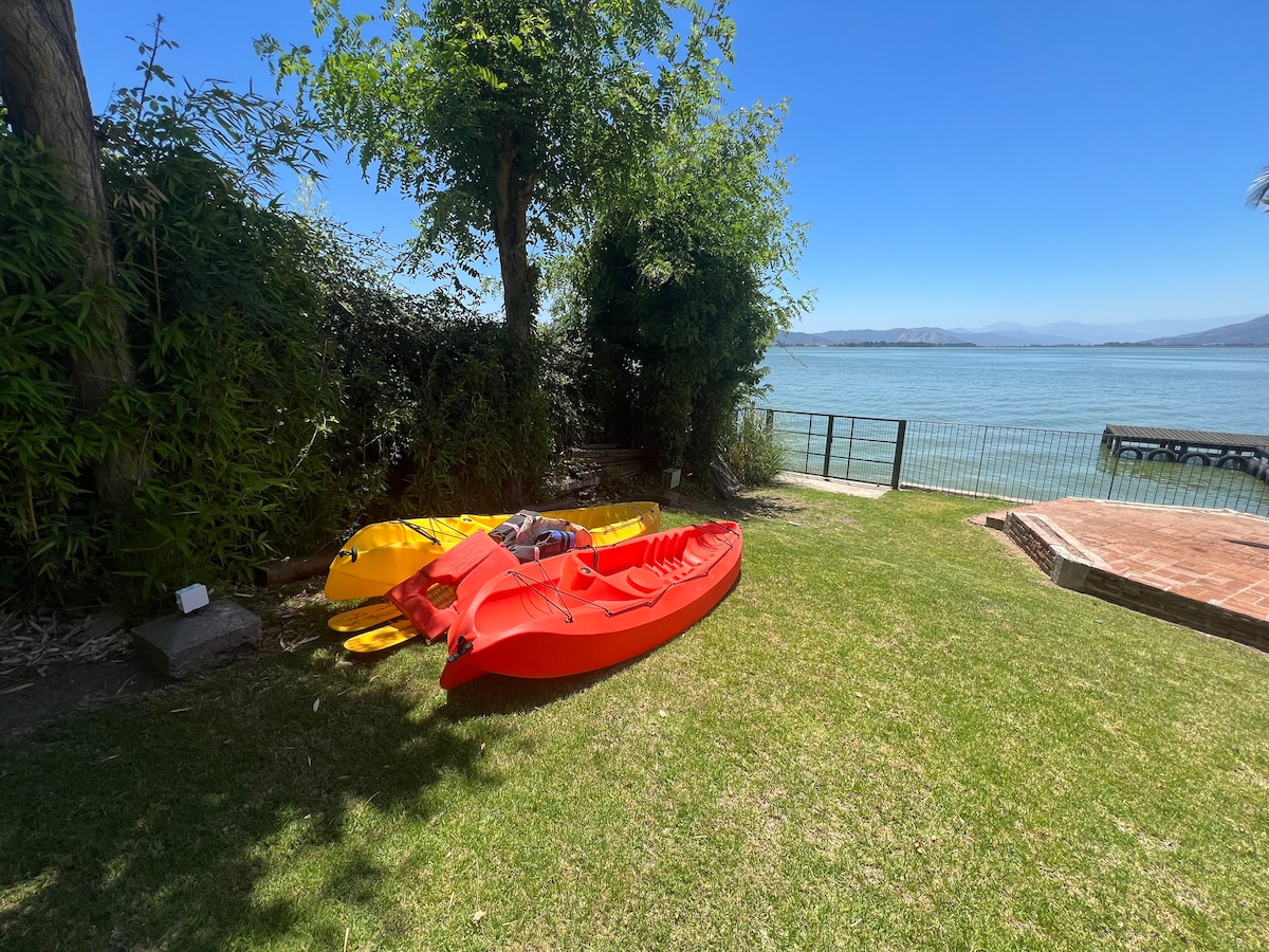 Two kayaks, one yellow and one red, rest on a green lawn near the water's edge. Lush greenery surrounds the area, providing shade and a sense of privacy, while the lake glistens in the sunlight, creating a peaceful scene.