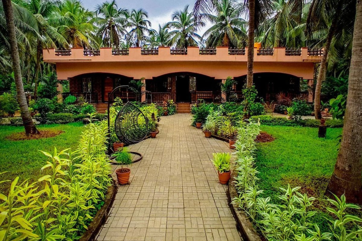A pathway lined with vibrant greenery leads to the spacious homestay. Potted plants adorn the sides, while palm trees provide shade. The structure features an elevated terrace with decorative railings, surrounded by lush gardens, creating a serene and inviting atmosphere.