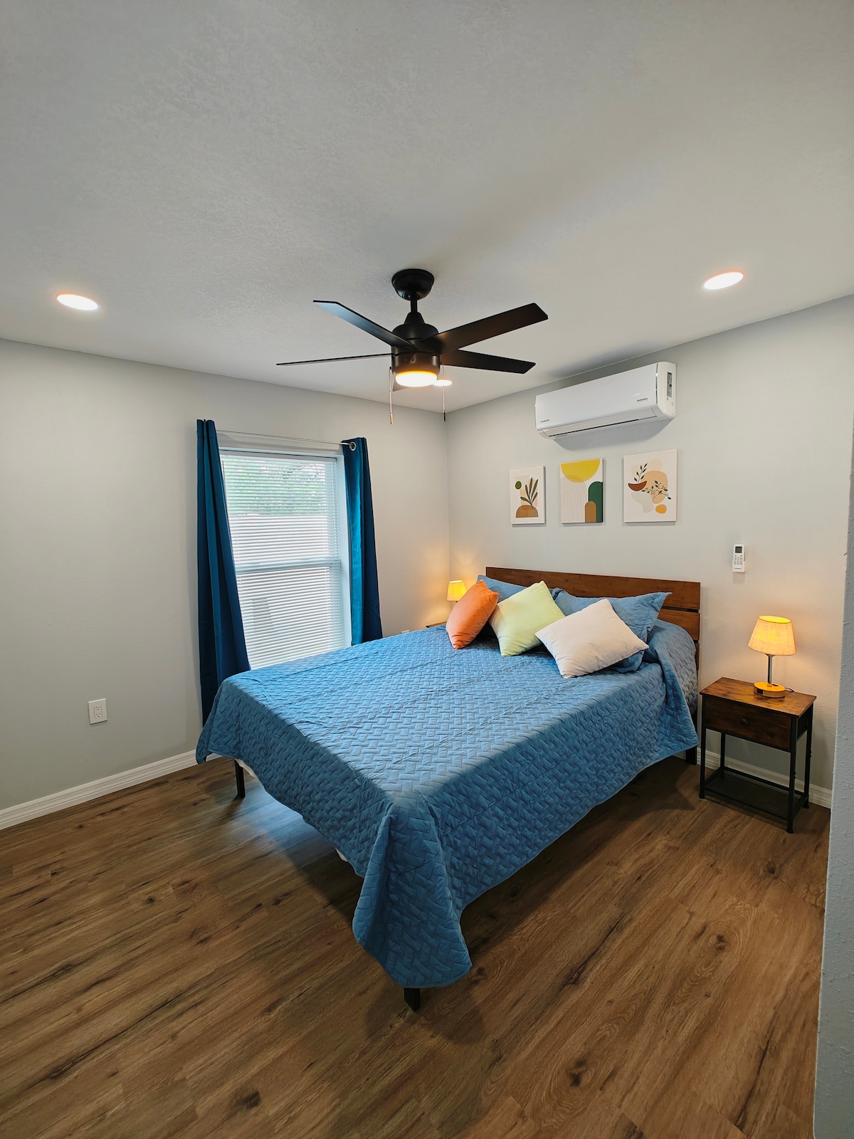 A bright bedroom features a spacious bed draped in a blue coverlet, accompanied by colorful decorative pillows. Natural light enters through a large window, adorned with blue curtains. A bedside table holds a lamp, and air conditioning controls are visible on the wall.