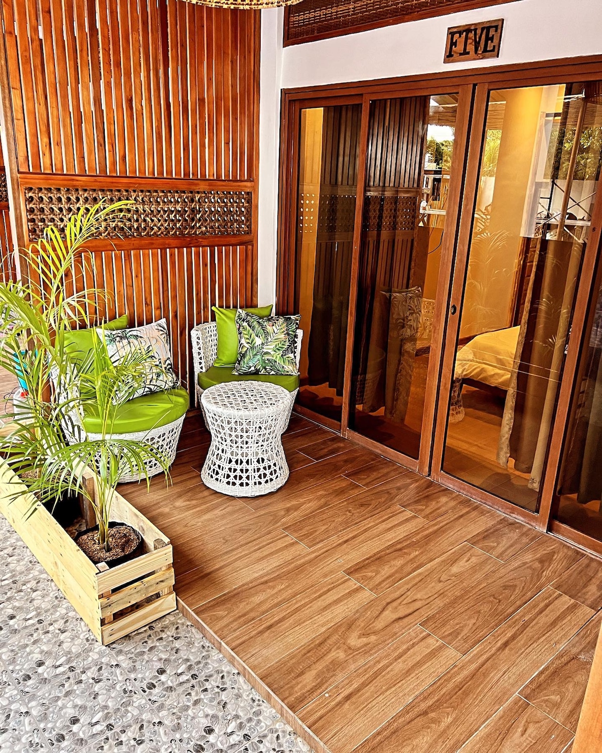 A cozy seating area is presented with two white wicker chairs adorned with green cushions. A small, round side table is positioned between the chairs. Natural light flows through sliding glass doors, while a potted plant adds a touch of greenery.