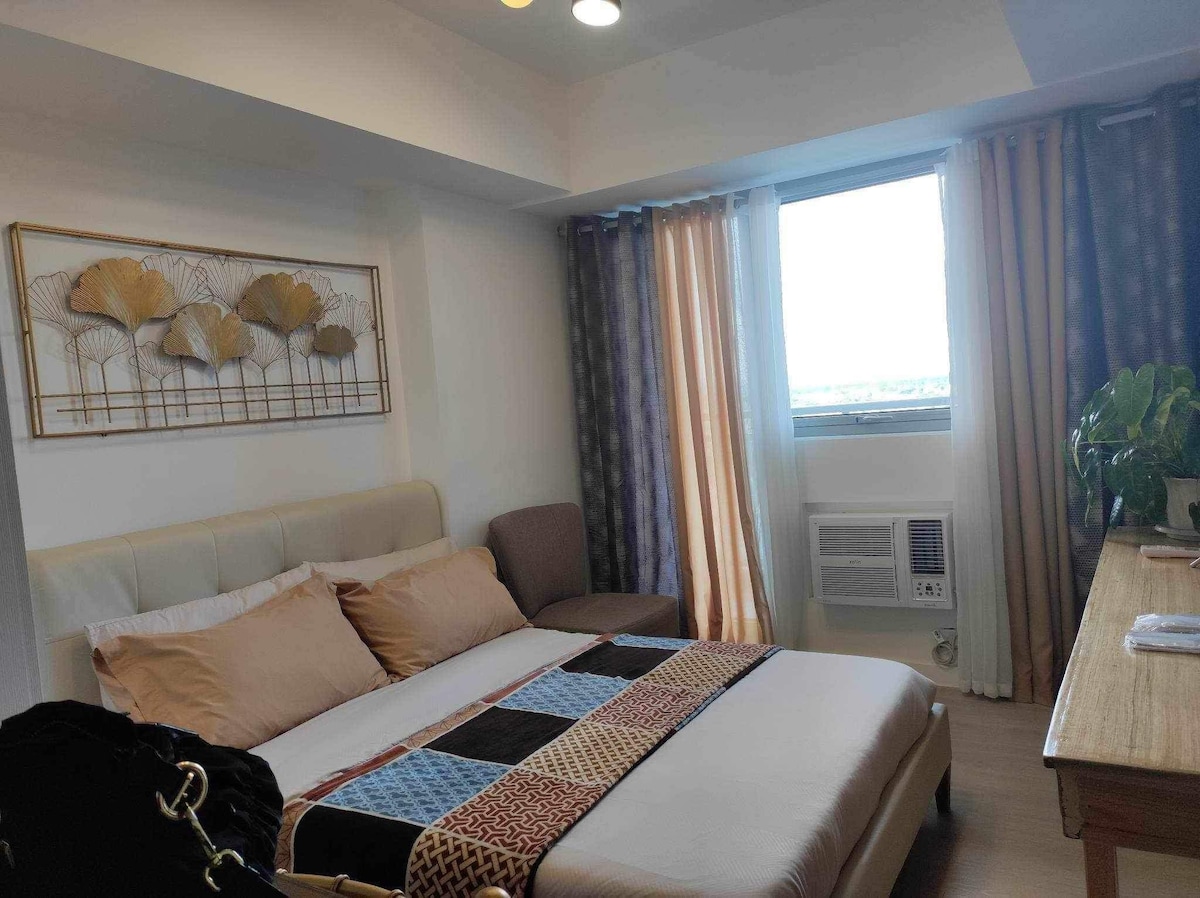 A cozy bedroom features a spacious bed adorned with neutral-toned bedding and a patterned throw. A single armchair is positioned near the window, which is framed by curtains in soft hues. A small plant is placed on a wooden dresser, contributing to the room's inviting feel.