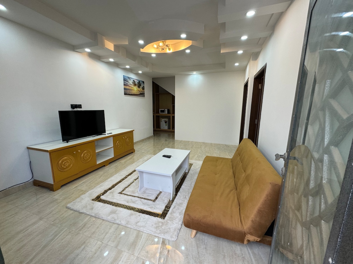 A bright living area features a brown sofa bed facing a white coffee table, positioned on a light-colored area rug. Soft lighting is provided by recessed ceiling lights, while a scenic landscape artwork adorns the wall above the television console.