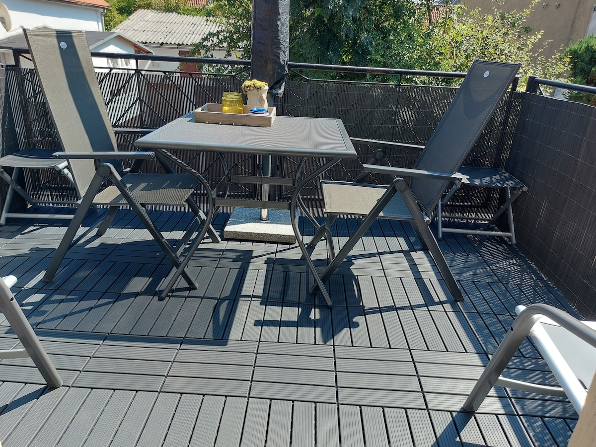 A spacious terrace is equipped with a table and four folding chairs, set on a composite floor. The area features greenery in the background, providing a peaceful outdoor setting, suitable for relaxation or casual dining.