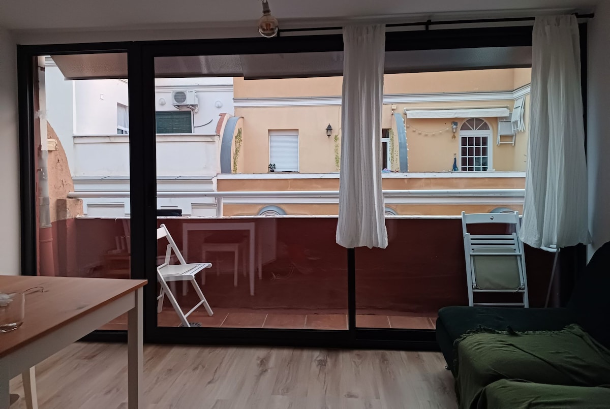 A large glass sliding door opens to a terrace, allowing natural light to fill the living area. Two white chairs are positioned on the terrace, with a small wooden table seen inside the space. Neutral tones dominate the decor, creating a calm atmosphere.