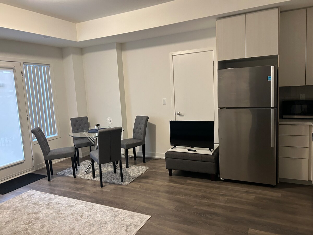A modern living area features an open space with a round dining table surrounded by four grey chairs. A sleek kitchen with stainless steel appliances, including a refrigerator, complements the design. A sofa is positioned near a TV stand against a wall, with a rug adding warmth.