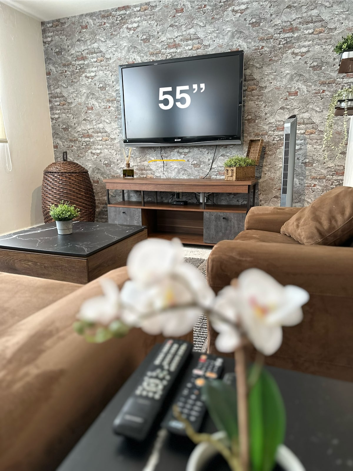 A comfortable seating area features a large television mounted on a textured stone wall. A coffee table sits in front of the seating, adorned with a potted orchid. Natural light gently fills the space, highlighting the warm tones of the furniture.
