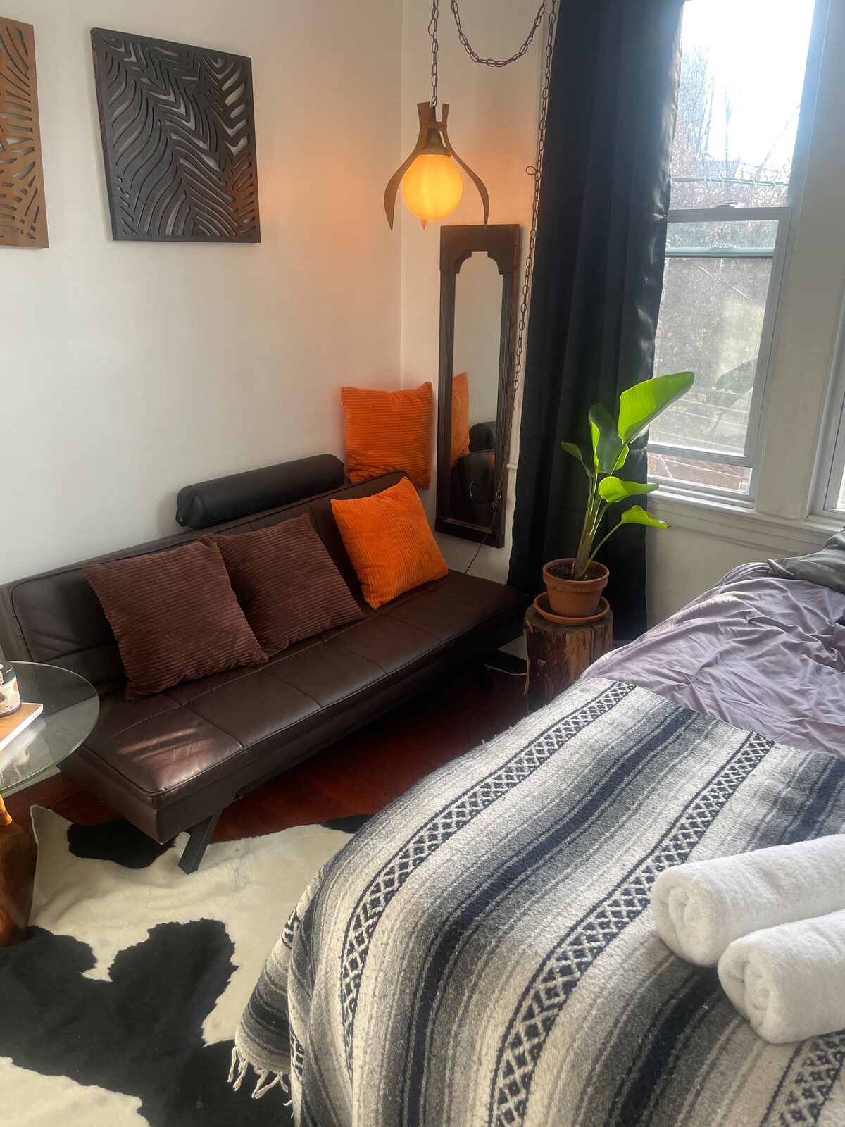 A cozy corner is highlighted by a stylish brown couch with two plush pillows, accompanied by a small glass table. A potted plant stands nearby, and a full-length mirror is mounted on the wall. Natural light filters through the large windows, illuminating the space.