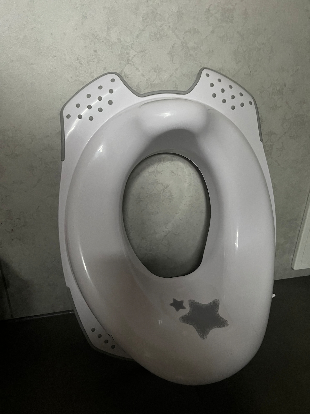 A child-sized toilet seat is shown, featuring a light gray color with star-shaped accents. The design includes small holes near the top for easy handling. The seat is positioned on a textured wall, reflecting a playful touch for children's use.