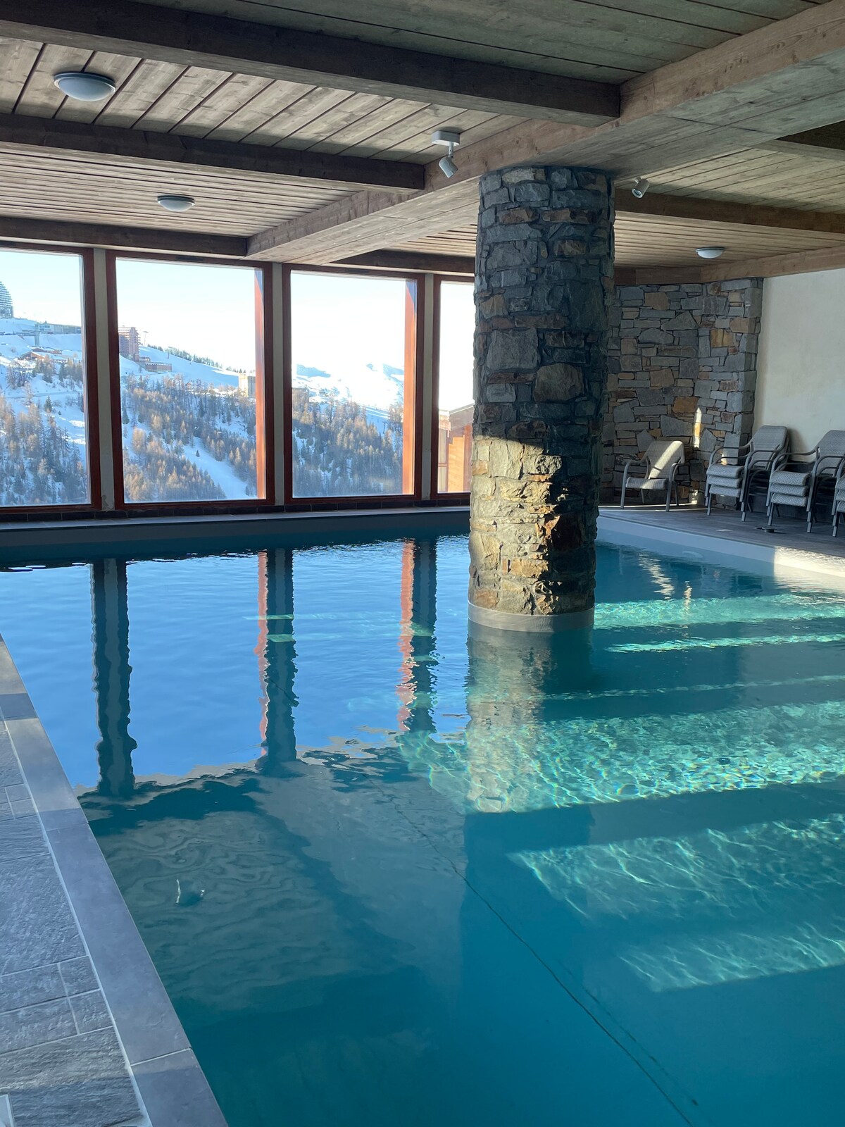 An indoor swimming pool is featured, surrounded by large windows that offer mountain views. Natural light reflects on the water's surface, enhancing the serene atmosphere. A stone column adds a structural element, while lounge chairs are positioned nearby for relaxation.