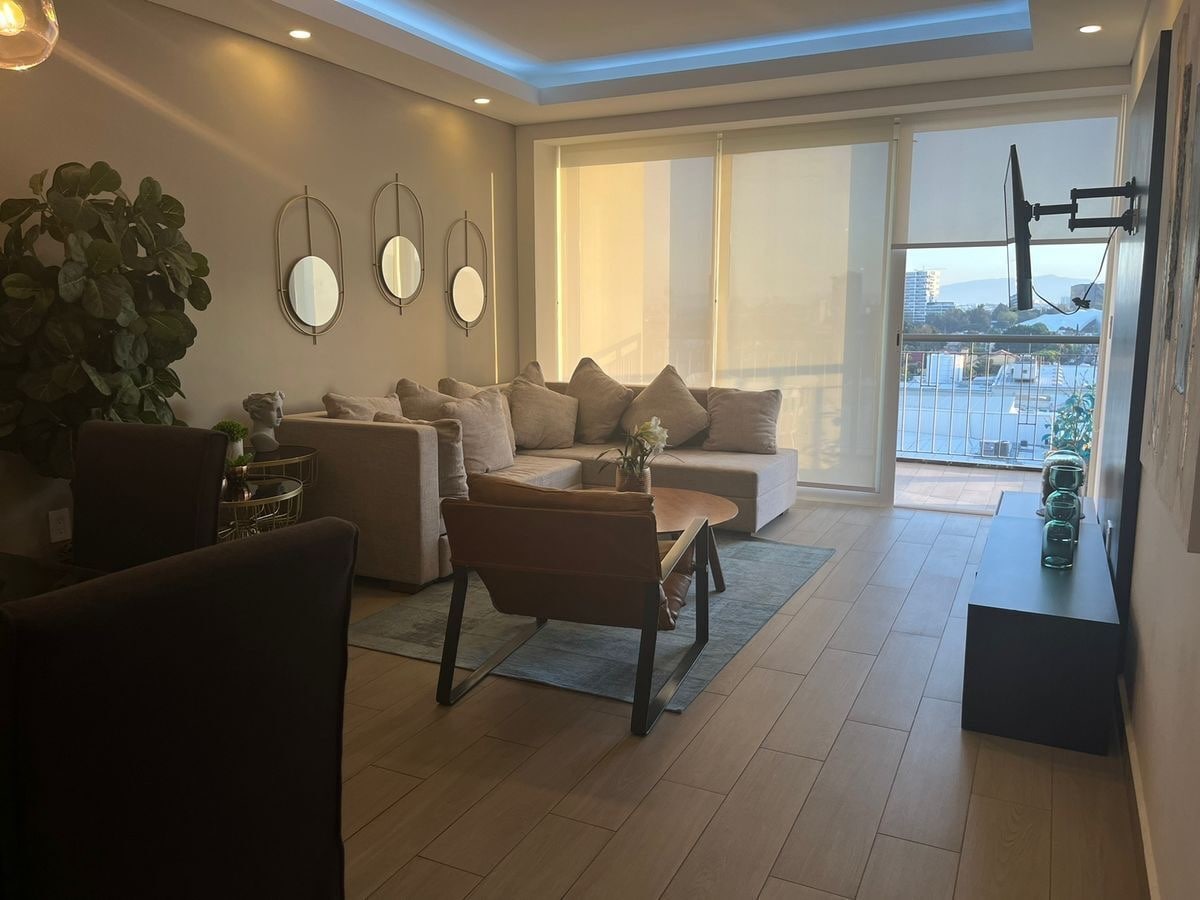 A modern living area is featured, showcasing a sectional sofa adorned with neutral cushions. A wooden coffee table is positioned centrally, complemented by a tasteful area rug. Large windows allow natural light to fill the room, accompanied by a flat-screen TV mounted on the wall.