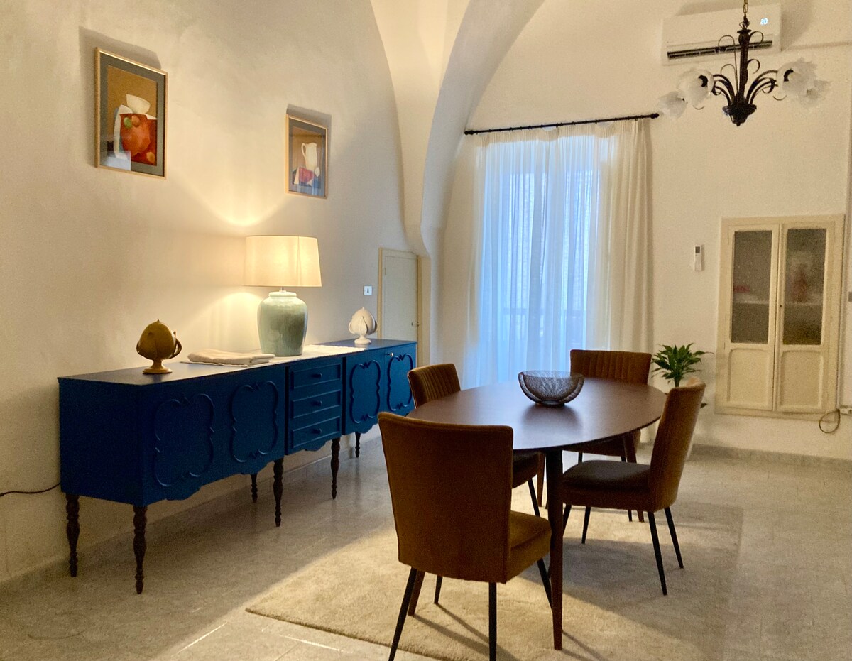 A spacious dining area features a round table surrounded by four upholstered chairs. A blue sideboard with decorative items is positioned against a light wall. A large window, dressed with sheer curtains, allows natural light to illuminate the space, complemented by a stylish chandelier.