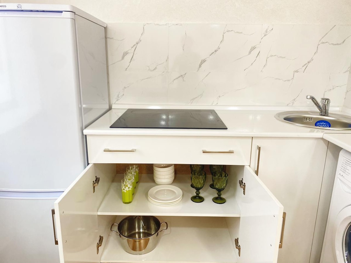 A modern kitchen displays white cabinetry with a pull-out drawer, revealing dinnerware and green glassware. A stainless steel pot rests inside the drawer. An electric cooktop and sink are visible along with a refrigerator, set against a marbled backsplash.