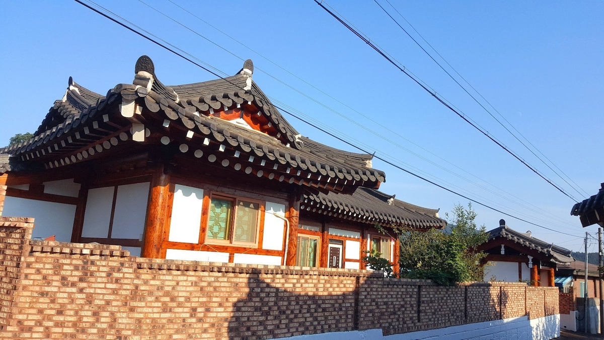 Traditional Korean houses are viewed from a distance, characterized by tiled roofs and wooden beams. A brick wall encloses the property, while a clear blue sky provides a serene backdrop. Lush greenery is visible, creating a harmonious blend with the architecture.