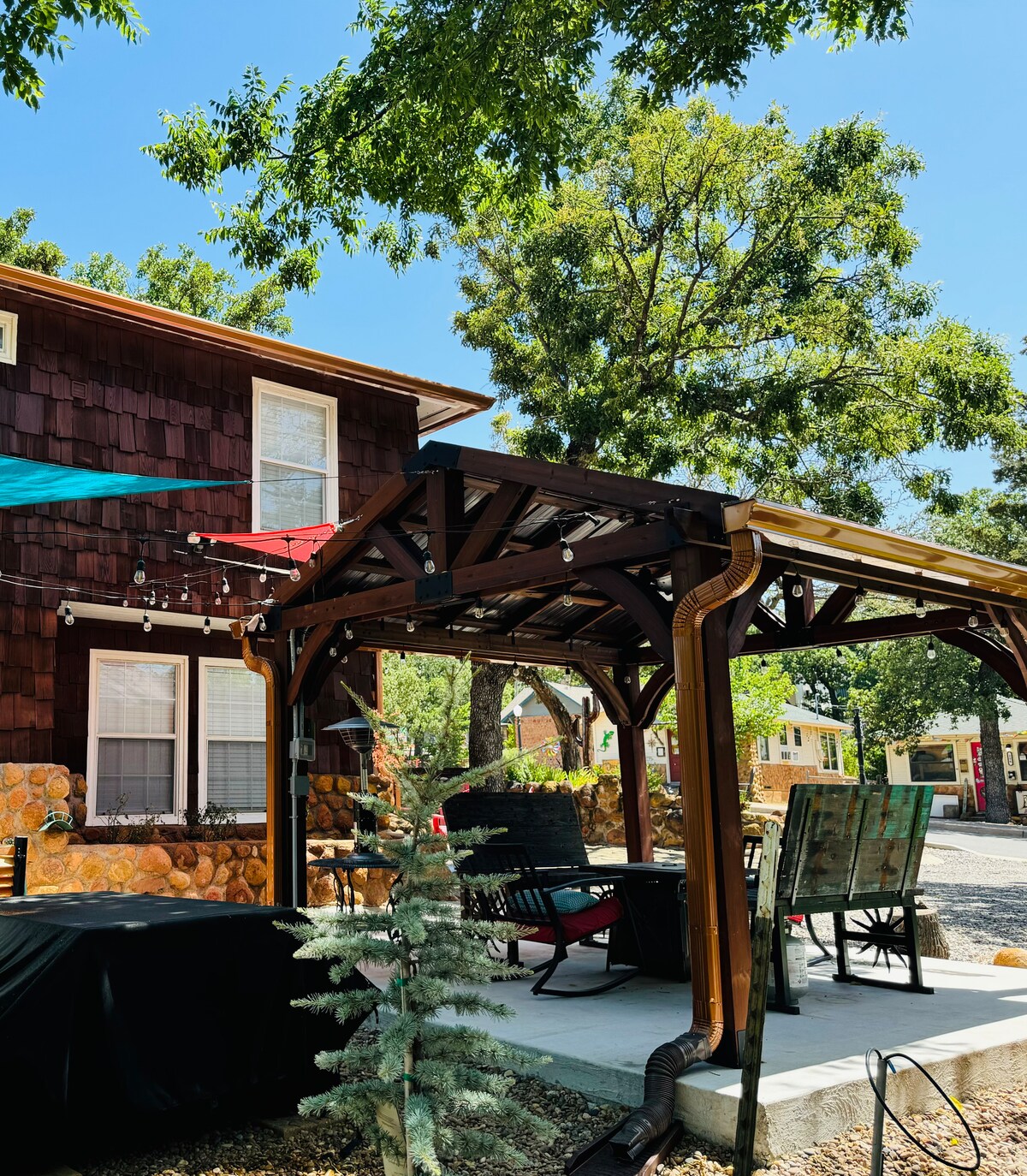 An extra-large covered patio features a wooden gazebo, with comfortable seating including a couple of chairs. Strands of lights hang gracefully above, and greenery surrounds the area, enhancing its inviting outdoor ambiance.