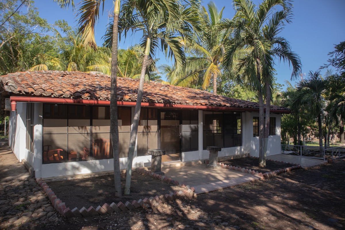 The exterior of the cabin is framed by lush palm trees, featuring a traditional tiled roof. The front porch includes several seating options and stone tables, providing a comfortable outdoor space. The surrounding area is landscaped with natural elements, enhancing the tranquil setting.