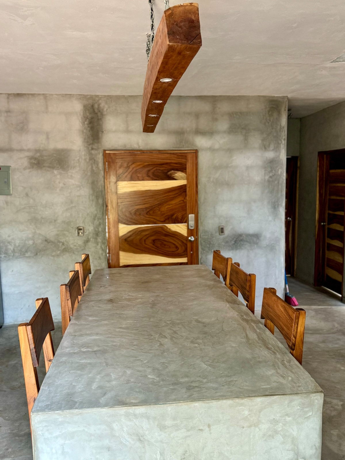 A spacious dining area features a long concrete table surrounded by wooden chairs. A wooden light fixture hangs above, illuminating the minimalist space. A stylish door with unique wooden panels is visible in the background, complementing the modern design elements.