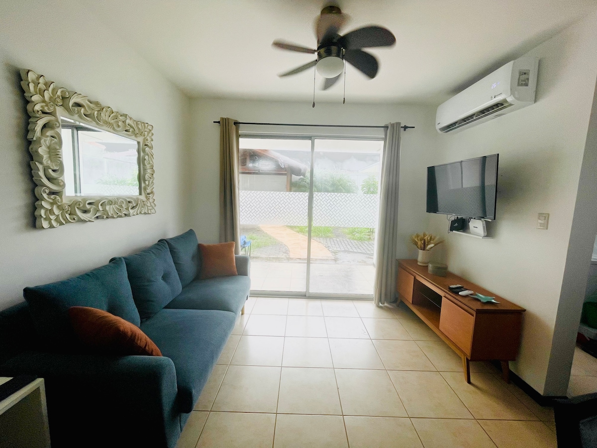 A living area features a comfortable blue sofa with accent pillows and a large mirror on the wall. A wall-mounted television is positioned near a wooden entertainment unit. Natural light flows through sliding glass doors, providing a view of the outdoor space.