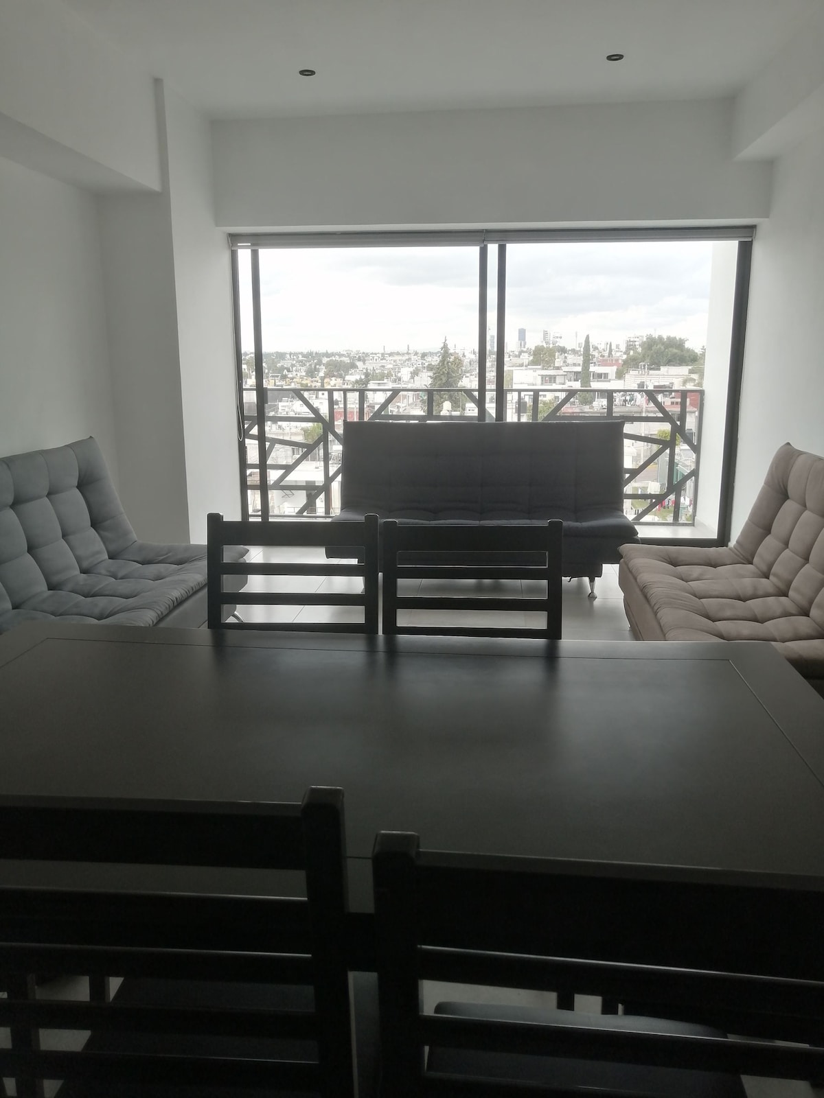 A bright living area features large windows offering a view of the cityscape. Two sofas in neutral tones sit on opposite sides of the room, while a dark dining table is set with several chairs, creating an inviting space for relaxation or meals.