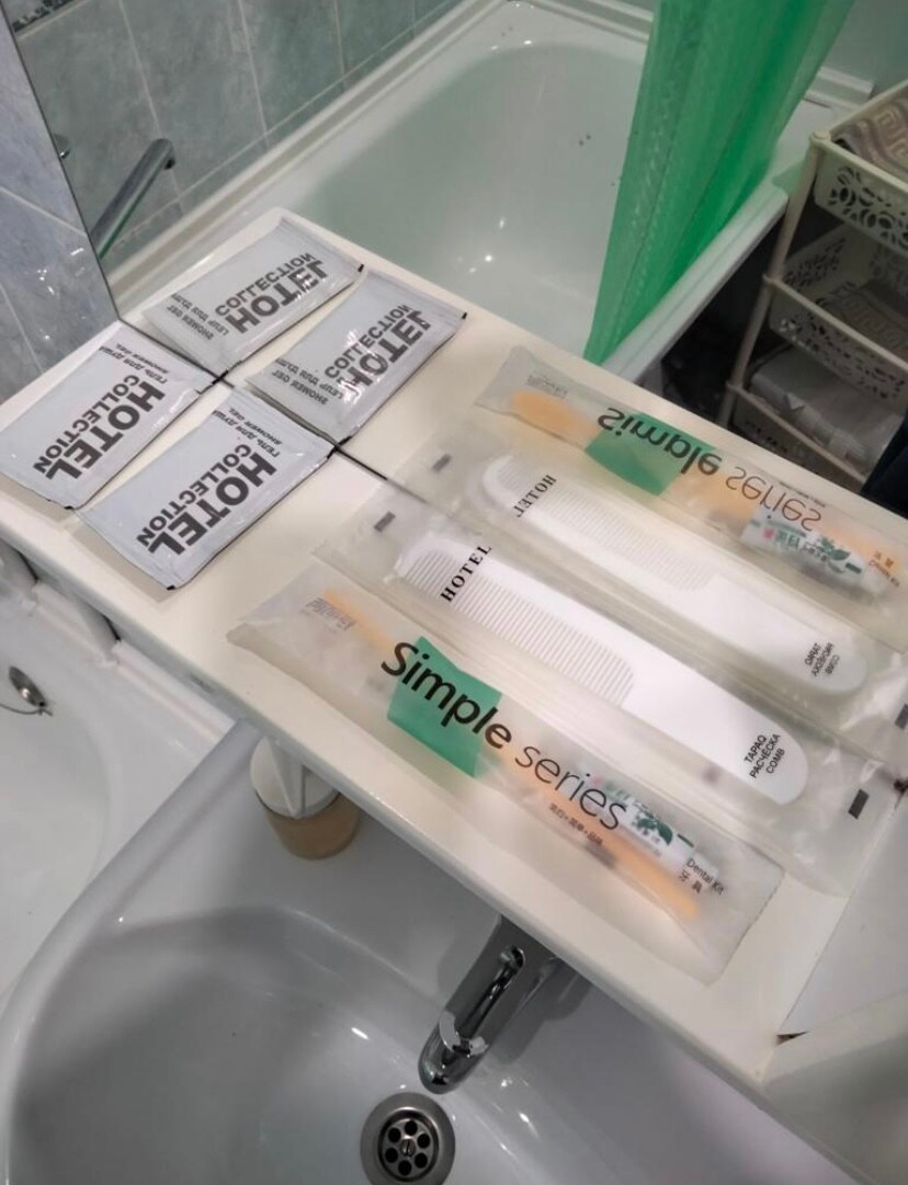 A bathroom countertop displays various toiletries, including a pack of toiletries labeled 'Simple Series'. A comb and two travel-sized items are arranged neatly beside multiple sealed packets labeled 'Hotel Collection'. The sink and a green shower curtain are visible in the background.