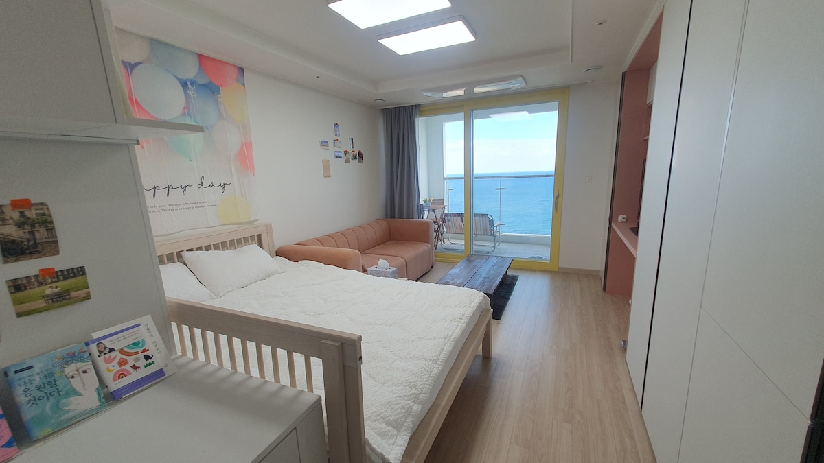 The room features a spacious bed positioned beside a large window offering views of the ocean. A cozy sofa in a warm shade is placed opposite the bed, with natural light illuminating the area. The soft color palette and decorative wall enhance the overall atmosphere.