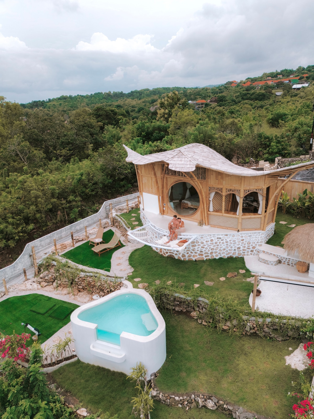 An aerial view of the bamboo house showcases its unique architecture surrounded by lush greenery. A small, inviting pool is seen alongside a landscaped area featuring grass and stone pathways. The home’s design includes large windows for natural light, connecting the indoor and outdoor environments.