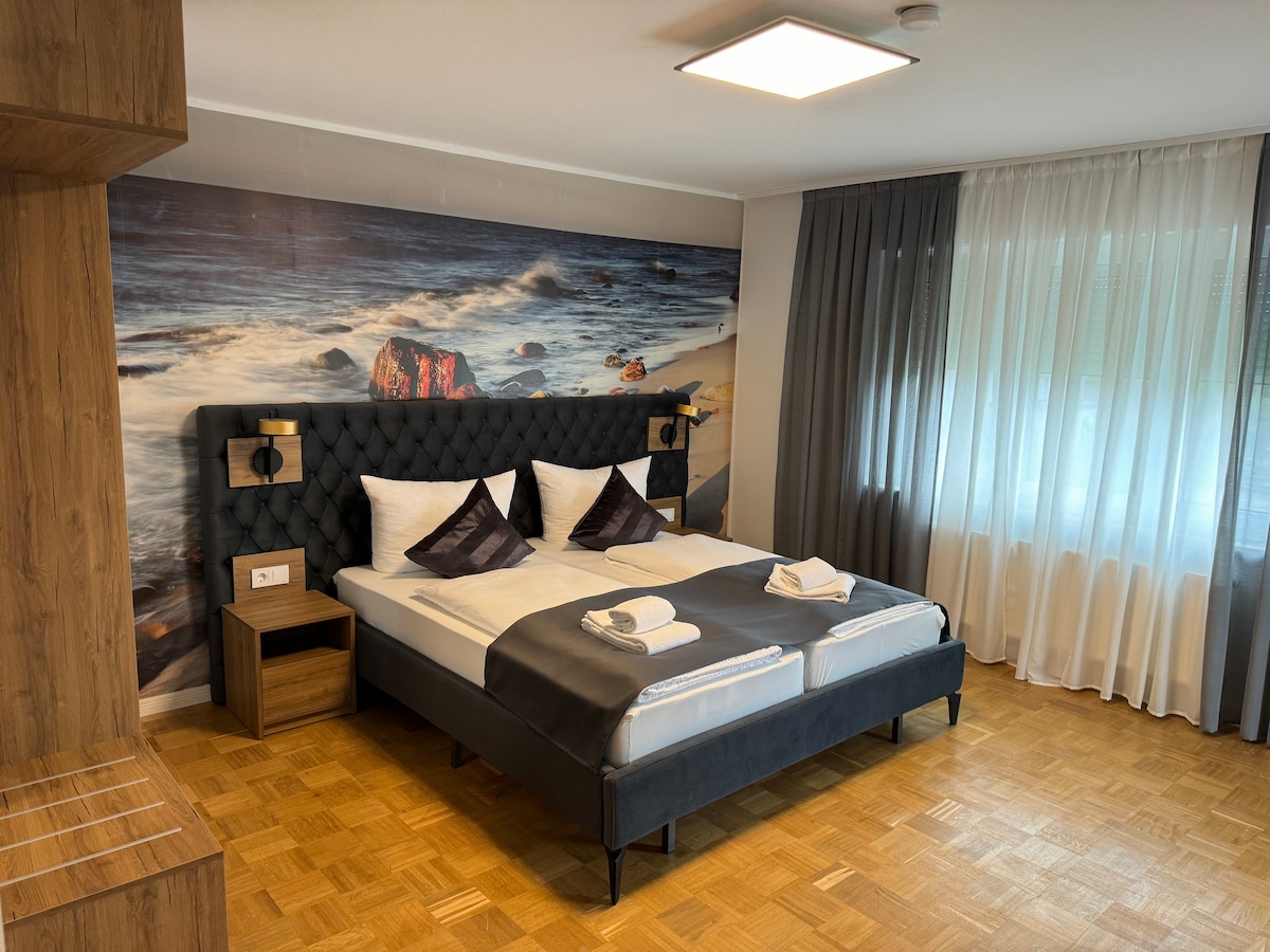 A modern sleeping area features a large, tufted bed with decorative pillows and fresh towels. A wall mural depicting a coastal scene adds to the decor. Natural light filters through sheer curtains, complementing the wooden elements of the room.