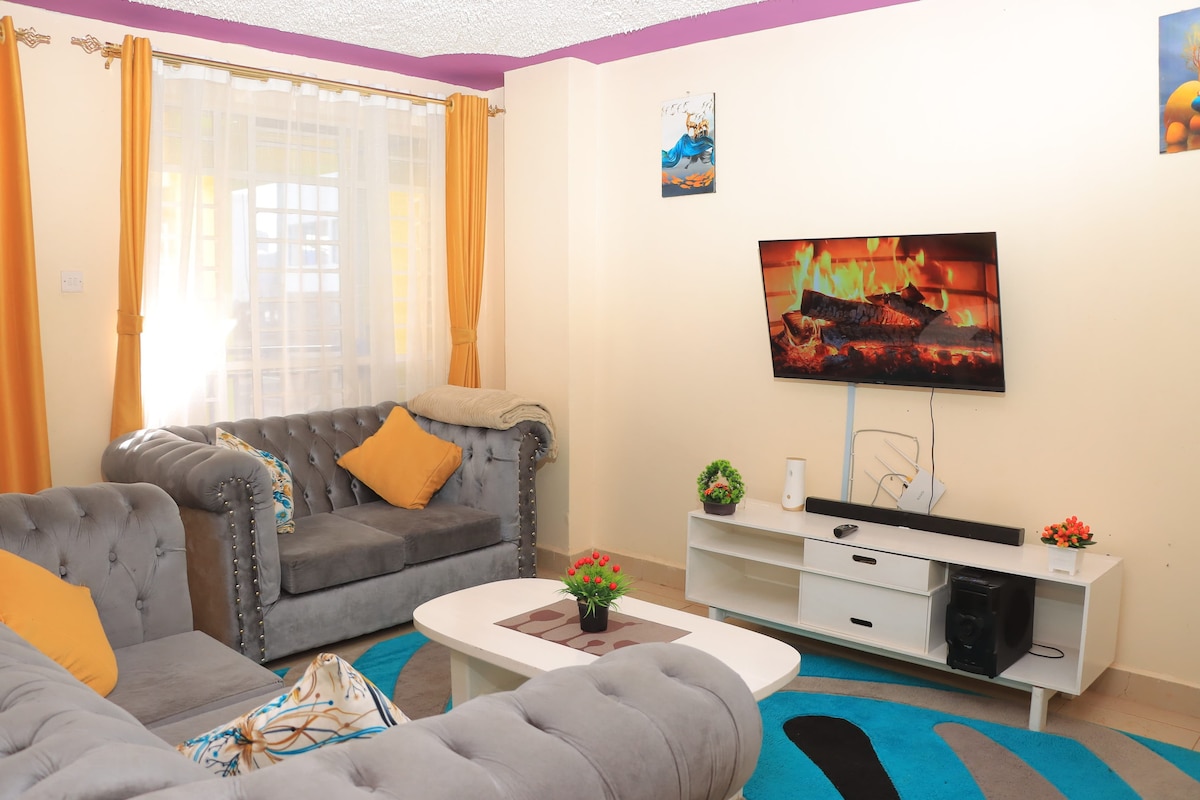 A cozy living area is featured, with a gray tufted sofa and a matching chair. A white entertainment unit displays a television, and vibrant cushions add color. The space is illuminated by natural light, with sheer curtains and wall art enhancing the inviting atmosphere.