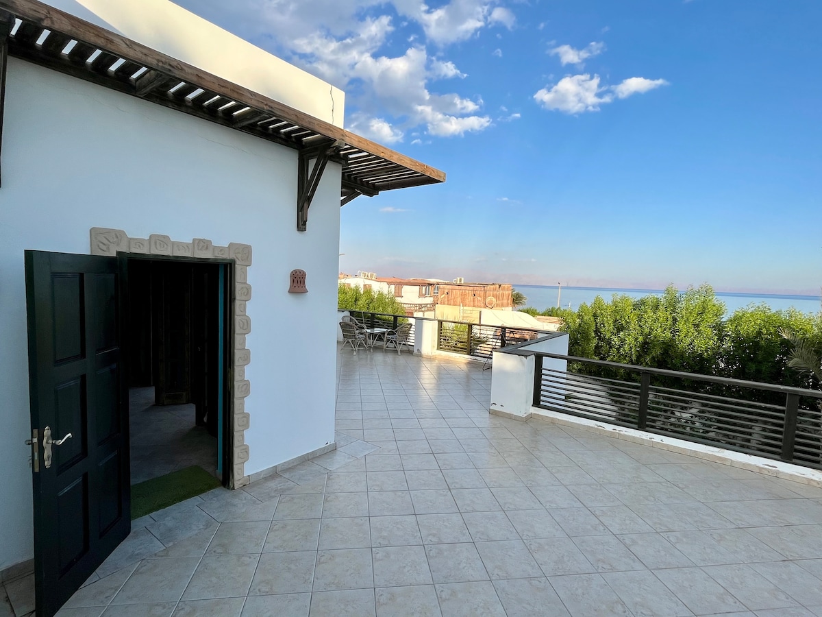 A large balcony is shown, featuring a clear view of the sea and mountains. The open door leads to a studio interior, while the tiled floor reflects natural light. A seating area is visible in the distance, surrounded by greenery.