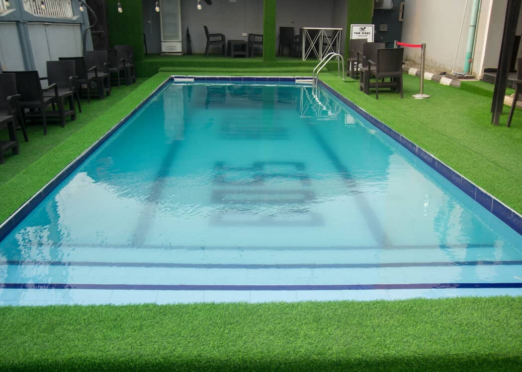 A communal swimming pool is surrounded by artificial grass and features clear blue water. Dark outdoor seating is arranged nearby, providing a space for relaxation. The area is well-maintained, enhancing a refreshing atmosphere for guests.