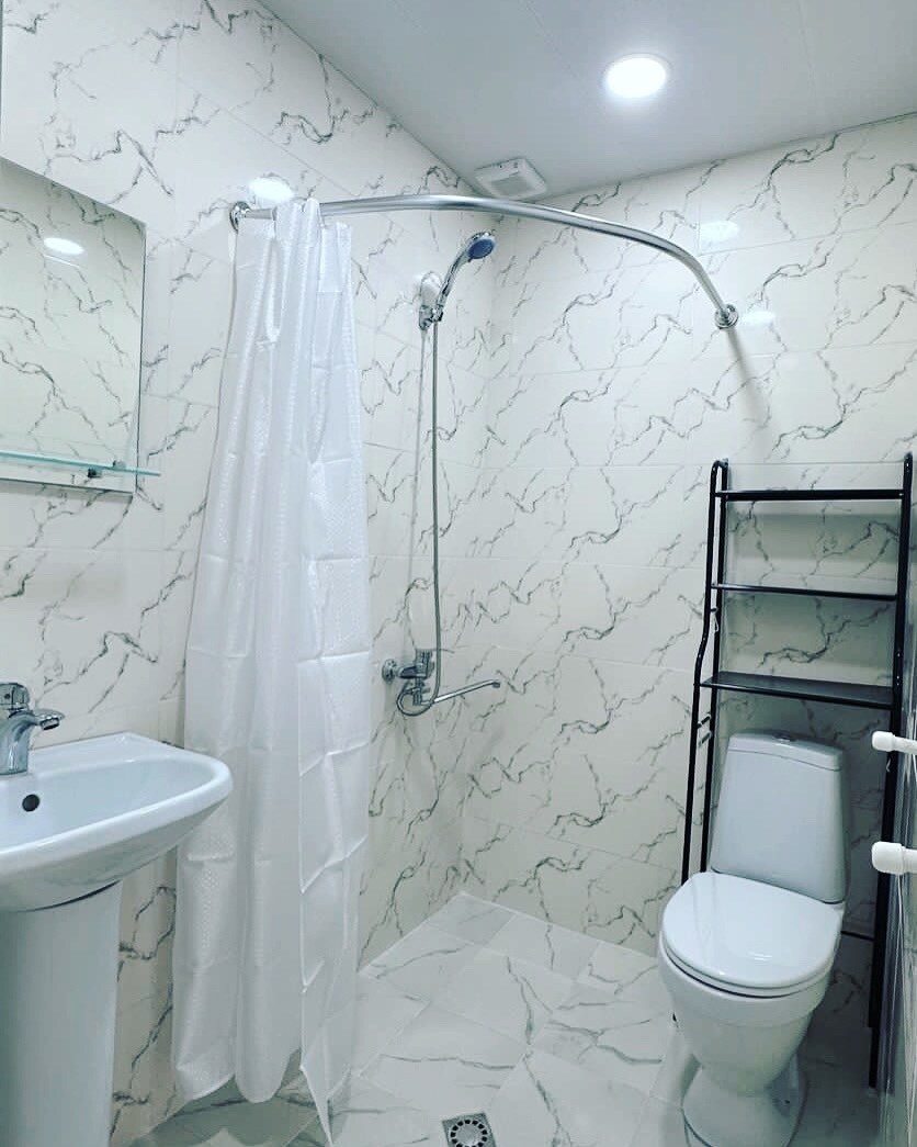 A modern bathroom is showcased, featuring a clean and minimalistic design. The space includes a walk-in shower with a transparent curtain, a sleek sink with a mirror above, and a toilet. Light-colored marble walls enhance the bright atmosphere.