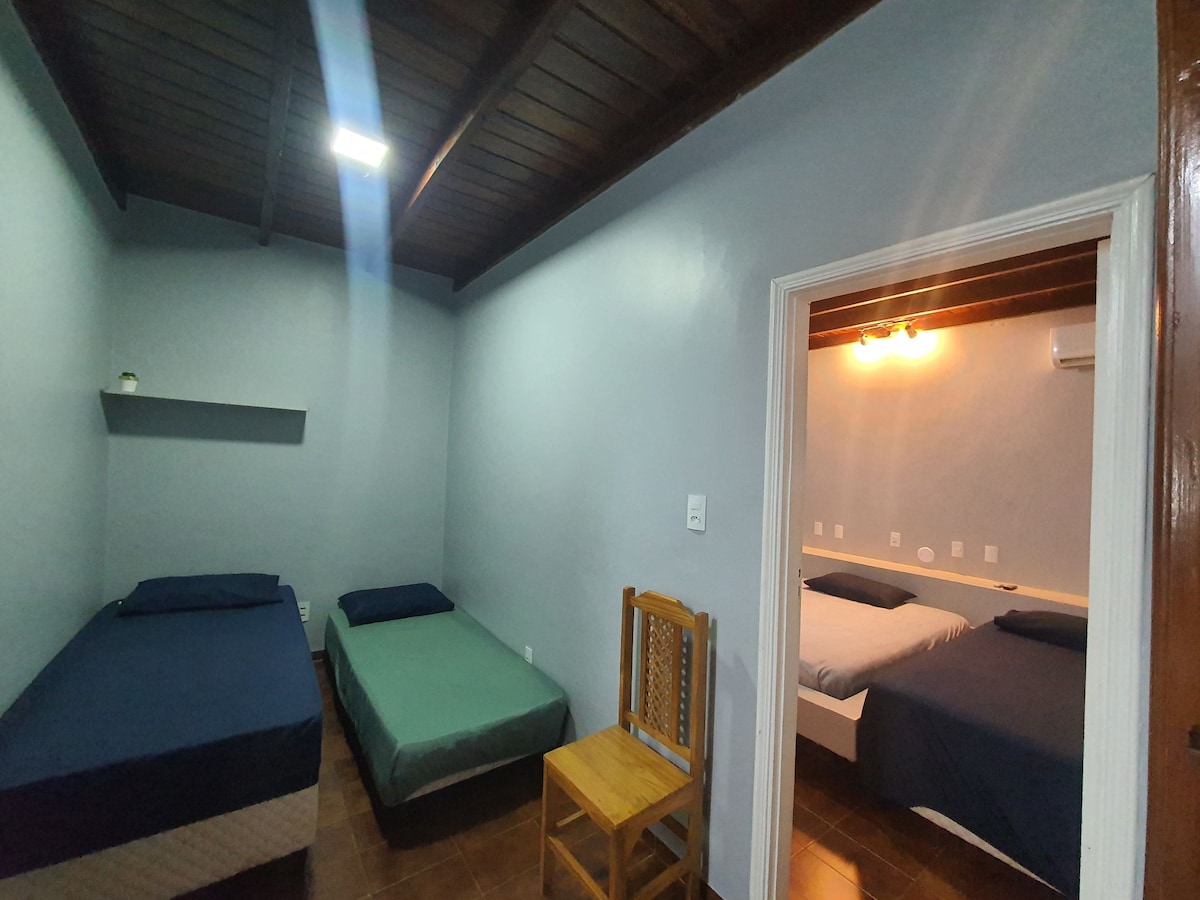A shared sleeping area is presented with two beds featuring blue and green bedding. A wooden chair is positioned nearby. A doorway leads to a second sleeping space with additional bedding arrangements visible in the background.