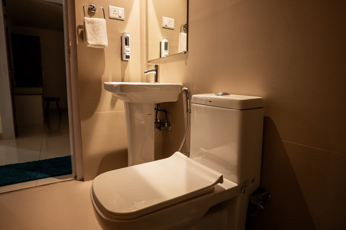 A clean bathroom is featured, showcasing modern fixtures. A white toilet and sink are positioned against smooth beige tiles, with a large mirror reflecting the space. Dispensers for soap and hand sanitizer are visible, enhancing accessibility and hygiene.