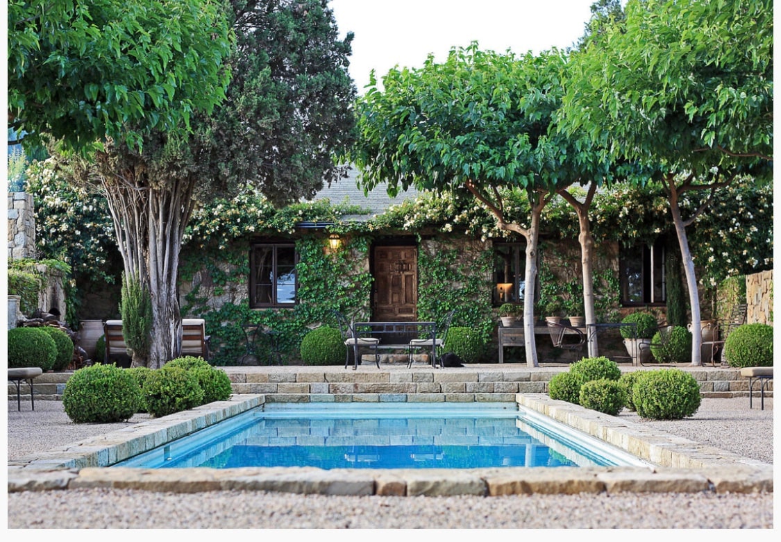 A serene courtyard is surrounded by lush greenery, featuring carefully shaped trees and manicured bushes. A crystal-clear pool reflects the surrounding landscape, while a stone pathway leads to the entrance of the rustic stone house, all framed by flowering vines.