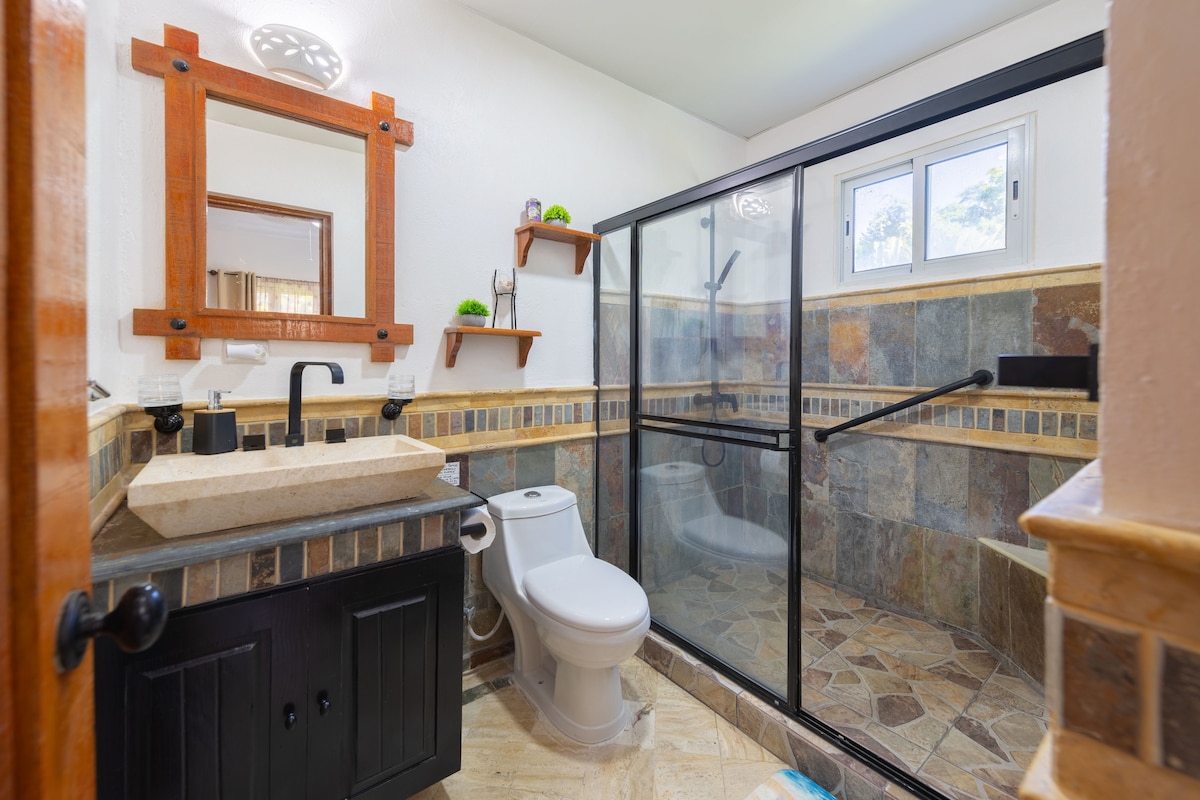 The bathroom features a modern design with a glass-enclosed shower and a natural stone countertop. Wooden shelves hold decorative items above the sink area, while textured tiles create a pleasing visual contrast. A small window provides natural light to the space.