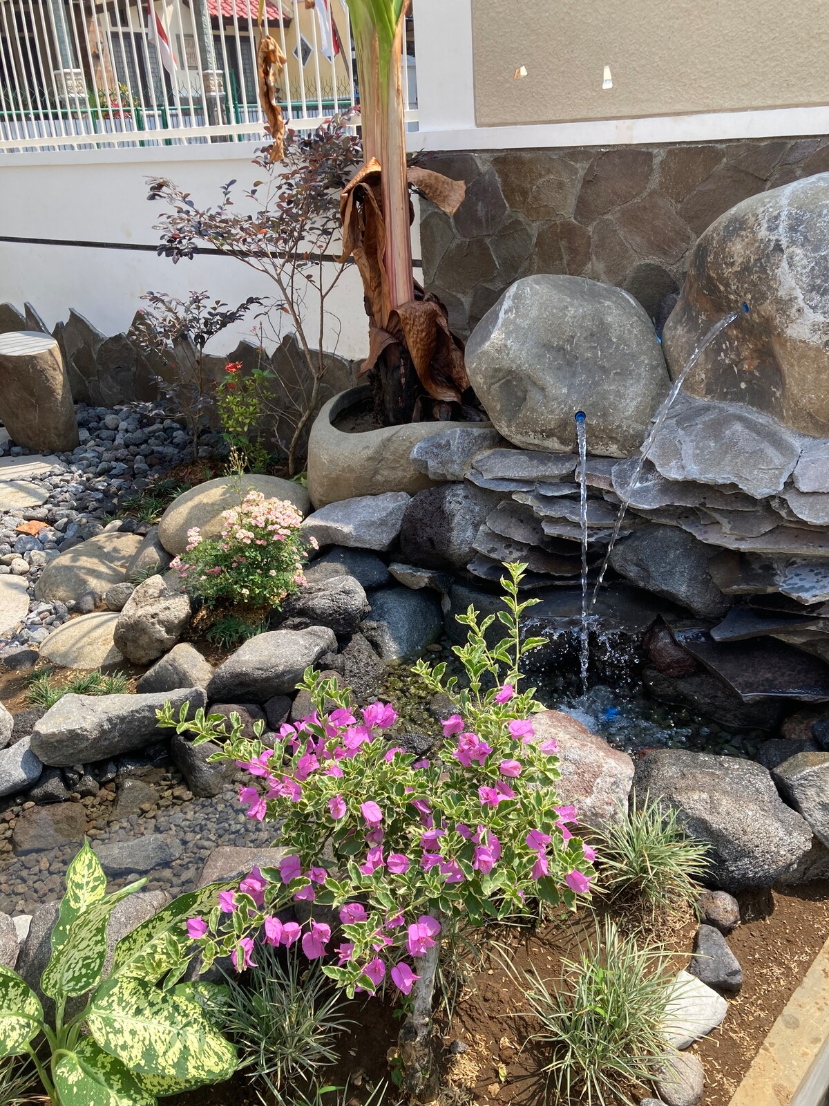 A serene garden area features a water fountain flowing gently over layered rocks. Colorful flowers, including purple bougainvillea and smaller blooms, are interspersed among carefully arranged stones, creating a tranquil natural setting.