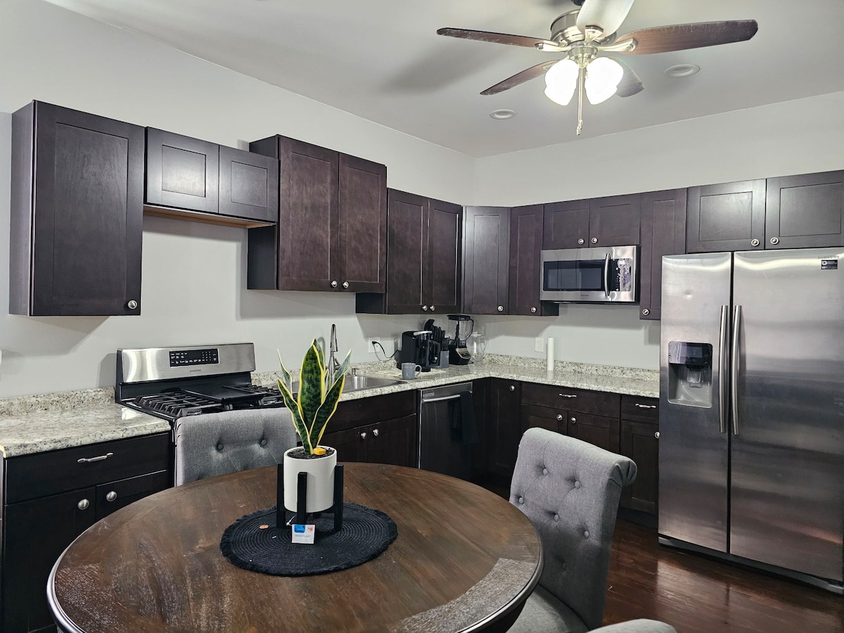 A modern kitchen features dark wooden cabinetry and stainless steel appliances, including a microwave and refrigerator. A circular dining table is accompanied by two upholstered chairs. A small potted plant is placed at the center of the table, adding a touch of greenery to the space.